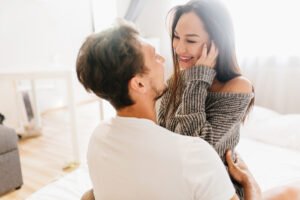 Short haired happy man embracing laughing young woman in gray soft clothes. Indoor portrait of romantic married couple enjoying morning in weekend together..